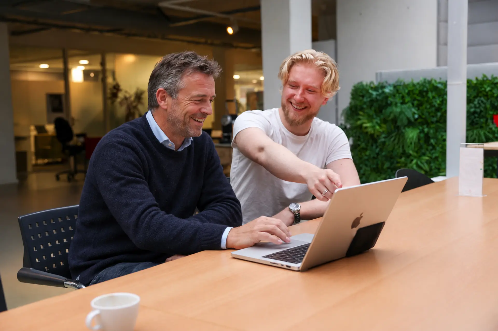 Twee mensen zitten aan een tafel met een open laptop; één van hen wijst naar het scherm